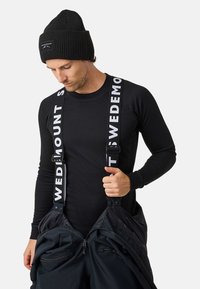 Zwarte geribbelde lange mouwen shirt gecombineerd met zwarte sneeuwbroeken met dikke witte banden. Muts met logo aan de voorkant. Zachte textuur, sportief ontwerp.