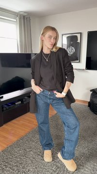 Persona con cabello rubio posa en interiores vistiendo blazer negro, camisa negra, jeans azules y zapatos beige, de pie sobre una alfombra gris cerca de un televisor y una obra de arte enmarcada.