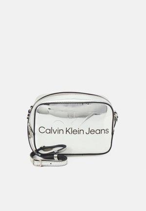Silver rectangular crossbody bag with a glossy finish, featuring "Calvin Klein Jeans" logo in black, a zip closure, and an adjustable strap.