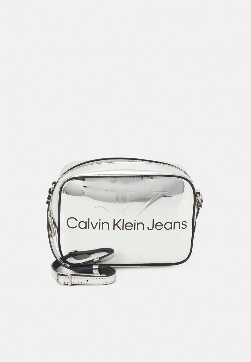 Silver rectangular crossbody bag with a glossy finish, featuring "Calvin Klein Jeans" logo in black, a zip closure, and an adjustable strap.
