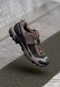 Brown and gray athletic shoe with a textured upper, rubber soles, and a quick-lace system, featuring a supportive design for outdoor activities.