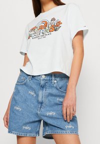 Žena v světle modrém zkráceném tričku s barevným květinovým potiskem "Tommy Jeans" a modrých denimových šortkách s výšivkou loga "Tommy Jeans".