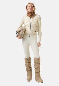 Cream puffer jacket with a high collar and zip, paired with cream fitted leggings. Chunky beige ski boots with gathered textile detail.