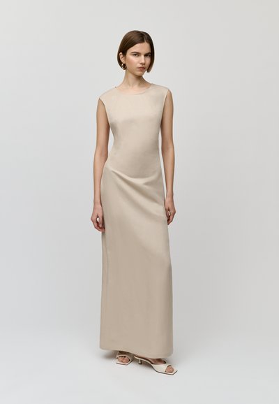 Robe maxi sans manches beige avec une texture lisse et satinée, dotée d'un col rond et d'un design minimaliste. Portée avec des talons ouverts.