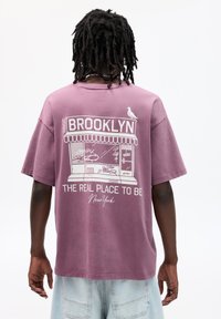 Fialové bavlnené tričko s grafikou obchodu a textom "BROOKLYN" a "THE REAL PLACE TO BE" v bielej farbe, vo voľnom strihu.