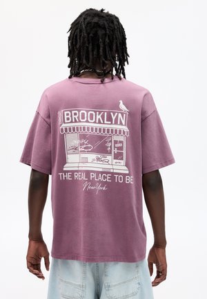 Tricou din bumbac violet cu un grafic al unei vitrine care include textul "BROOKLYN" și "LOCUL REAL UNDE SĂ FII" în alb, cu un croi lejer.