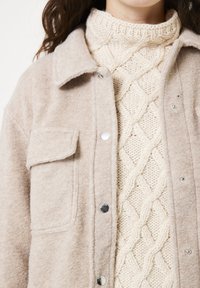 Chaqueta de camisa de franela beige con cierre de botones y bolsillo en el pecho, superpuesta a un suéter de punto trenzado en color crema. Apariencia de tejido suave.