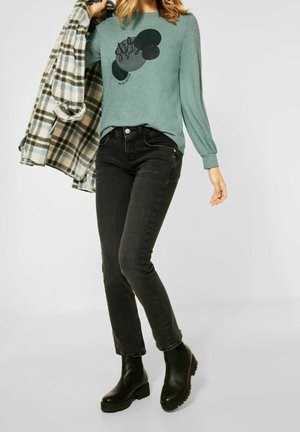 Vrouw draagt een turquoise lange mouwen shirt met abstracte print, zwarte spijkerbroek en zwarte laarzen, houdt een blauw-beige geruite jas over haar schouder.