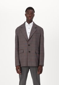 Rutete blazer i grå, oransje og blå. Har store slåer, to frontlommer og en knappelukking. Brukt over en hvit skjorte.