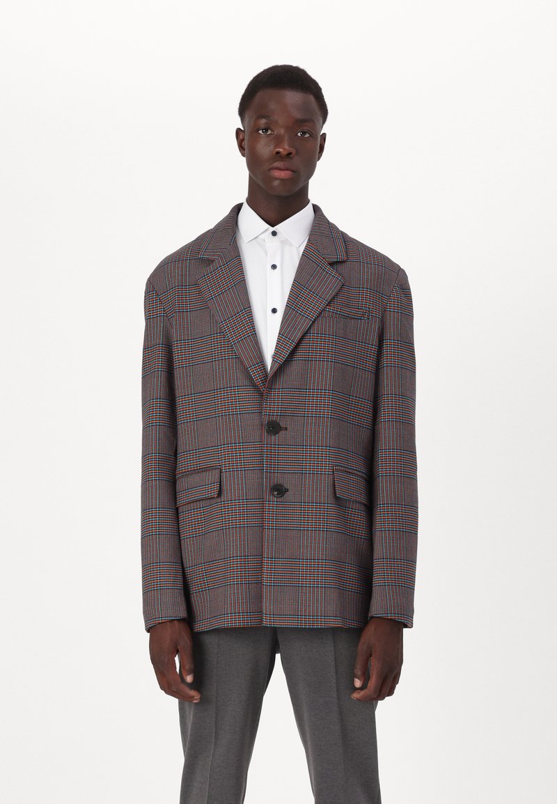 Rutete blazer i grå, oransje og blå. Har store slåer, to frontlommer og en knappelukking. Brukt over en hvit skjorte.