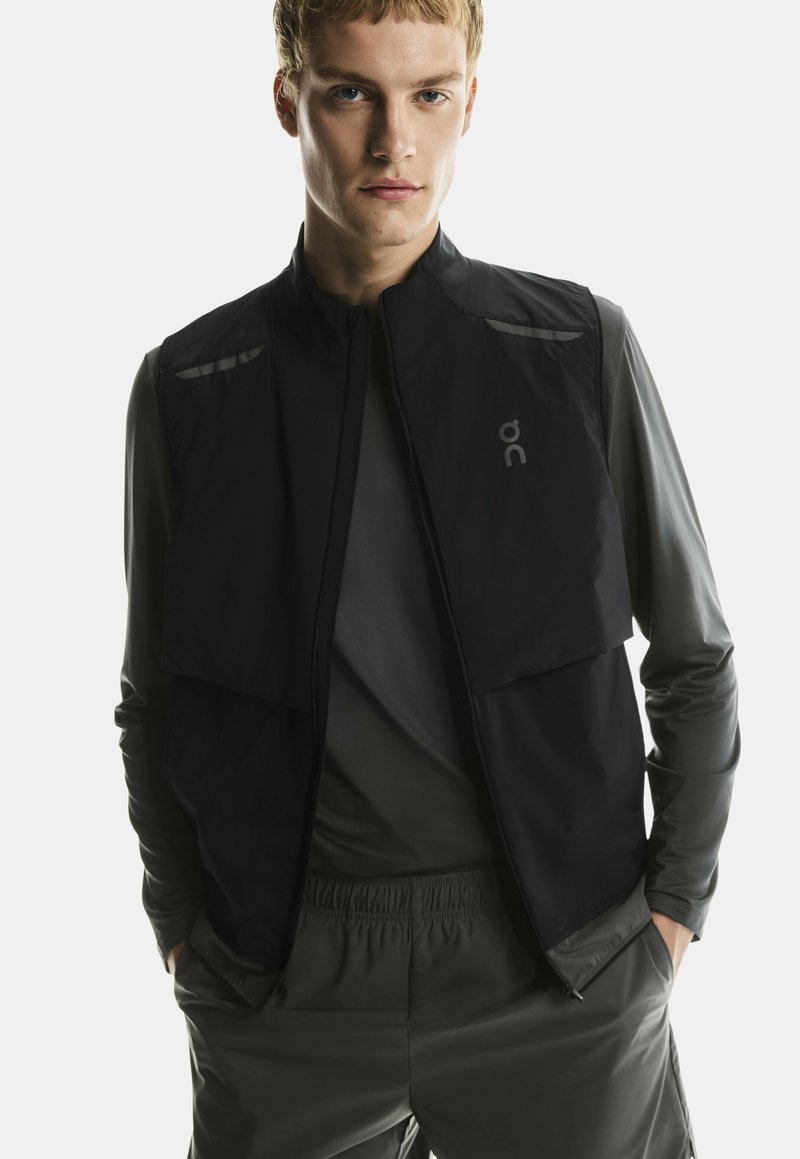 Jeune homme portant un gilet noir sans manches par-dessus une chemise à manches longues foncée et un pantalon assorti, les mains dans les poches, fond neutre.