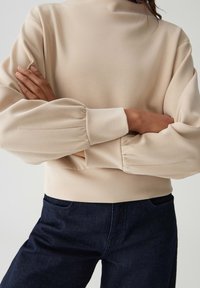 Felpa beige a collo alto con maniche lunghe a sbuffo e polsini a costine. Abbinata a jeans blu scuro, enfatizzando una texture liscia e un design aderente.