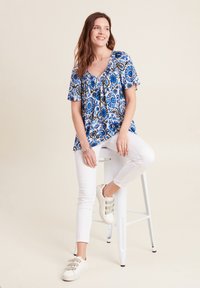 Blau-weißer gemusterter Oberteil mit kurzen Ärmeln, Rundhalsausschnitt und Raffung. Kombiniert mit weißen Slim-Fit-Hosen und beigen Sneakers.