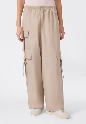 Beige Cargo-Hosen mit weitem Bein, mehreren Klappentaschen und Kordelzug-Details, getragen mit weißen Turnschuhen vor neutralem Hintergrund.