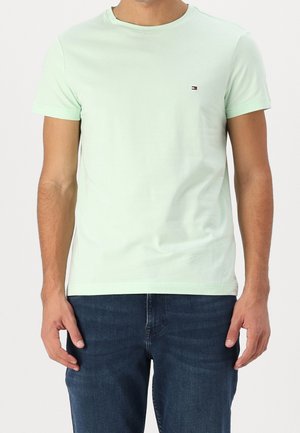Mężczyzna w jasnozielonym T-shircie z krótkim rękawem i małym logo na piersi oraz ciemnoniebieskich dżinsach, stoi z rękami swobodnie opuszczonymi wzdłuż ciała.