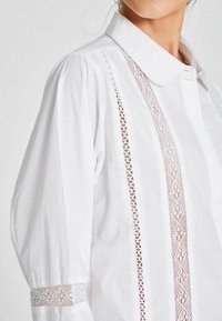 Valge puuvillane särk, millel on broderie anglaise aktsendid käistel ja esiosas, klassikaline krae ja kergelt paisutatud varrukad.