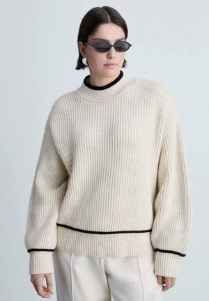 Pull en tricot côtelé crème avec des bandes noires aux poignets et à l'ourlet, présentant un col montant et une coupe décontractée, associé à des lunettes de soleil sombres.