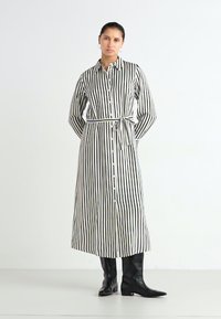 Šaty z čierno-bieleho pruhovaného materiálu s dlhými rukávmi, gombíkovým zapínaním vpredu, pásom v páse a dlhým lemom, skombinované s čiernymi vysokými čižmami po kolená.