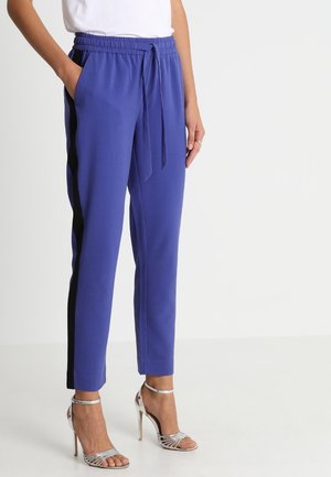 Kobieta ubrana w królewsko niebieskie spodnie z czarnymi bocznymi paskami, z wiązanym w talii sznurkiem, biała bluzka i srebrne sandały na wysokim obcasie, stojąca z rękami w kieszeniach.