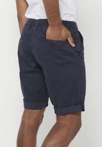 Man in opgerolde marineblauwe shorts en een wit shirt, met een hand gedeeltelijk in zijn achterzak, gezien van middenrug tot knieën.