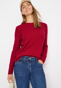 Czerwony sweter z prążkowanej dzianiny, z długimi rękawami, okrągłym dekoltem i luźnym krojem, zestawiony z niebieskimi dżinsami. Brak widocznych wzorów lub dodatków.