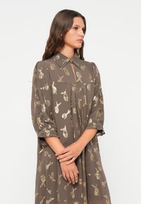 Ruskea mekko, jossa on kaulus, avainaukko-yksityiskohta ja lyhyet hihat. Sisältää kultaisia paisley-kuvioita ja vähäisen muotoilun mukavuutta varten.