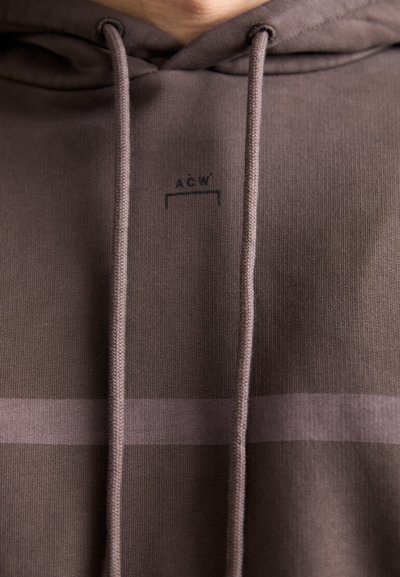 Sudadera marrón con bolsillo delantero, capucha y cordones. Incluye un logo sutil y un patrón de rayas horizontales en marrón más claro.