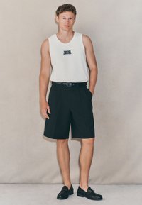 Canotta bianca senza maniche con testo ricamato in blu navy, abbinata a pantaloncini neri su misura e mocassini neri, con cintura e tasche laterali.