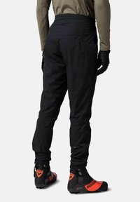 Schwarze Skihosen mit hoher Taille, die einen subtilen Glanz aufweisen, elastische Bündchen und eine Reißverschlusstasche besitzen. Kombiniert mit schwarz-roten Skischuhen.