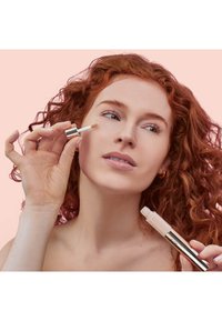 Concealer med applikator i form av en grävlingfot, silverlock, och genomskinlig kropp, som visas när den appliceras på kinden, mot en rosa bakgrund.