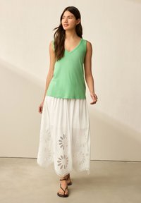 Top verde senza maniche con scollatura a V, abbinato a una gonna maxi bianca con design floreali ritagliati all'orlo e sandali neri.
