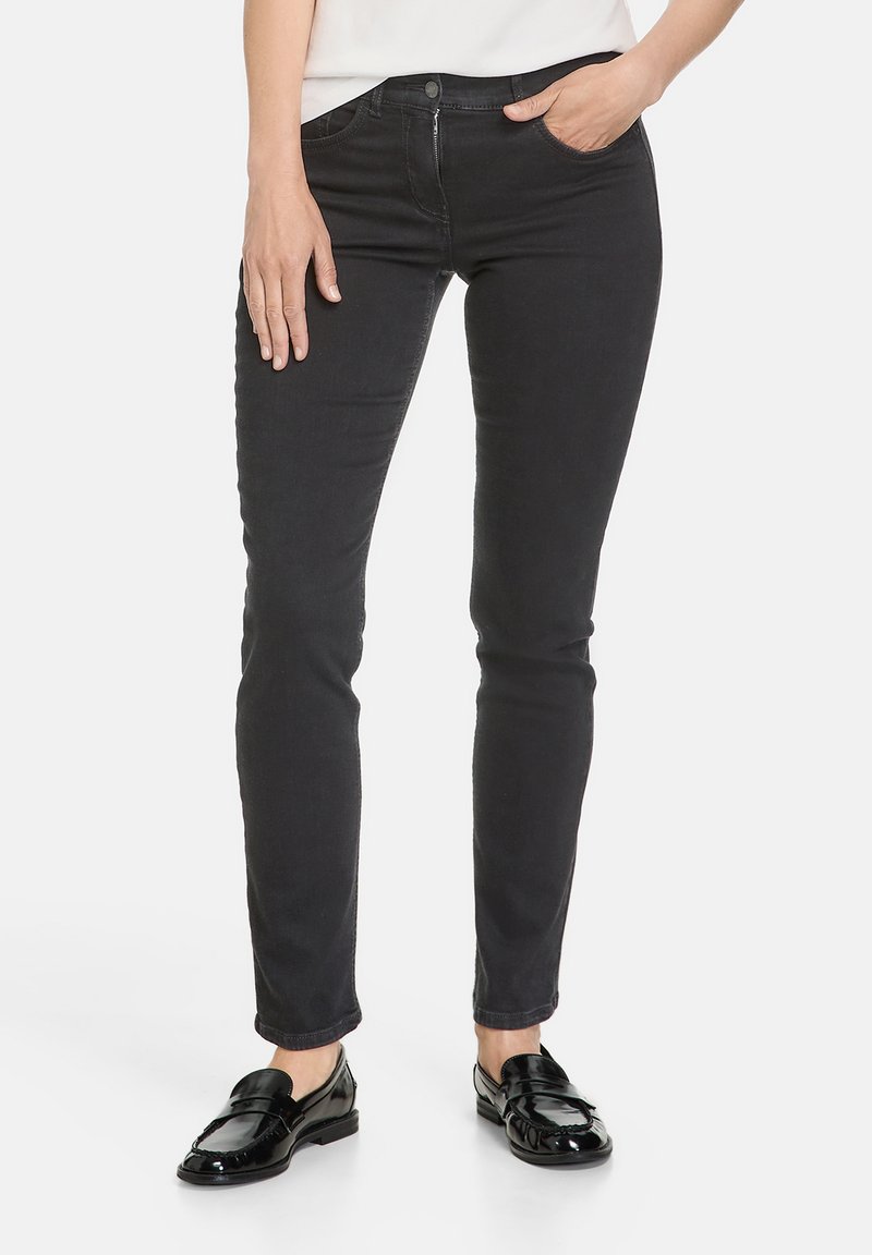 Schwarze, dehnbare Skinny-Jeans aus Denim mit Vorderfächern, Metallknopf und Reißverschluss sowie einer glatten, matten Textur. Getragen mit schwarzen Slippern.