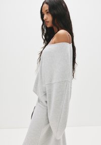 Sivá oversized mikina so širokými rukávmi, jemnou textúrou a minimálnym švom, nosená cez svetloružový top, zobrazená na neutralnom pozadí.