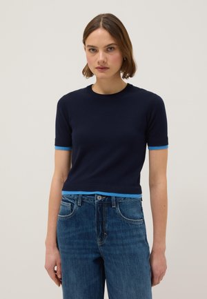 Vrouw draagt een nauwsluitende marineblauwe trui met korte mouwen en lichtblauwe bies, gecombineerd met een high-waisted blauwe jeans, staand tegen een effen achtergrond.