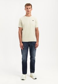Lichtgroen T-shirt met korte mouwen en een ronde halslijn, voorzien van een klein zwart logo, gecombineerd met donkerblauwe jeans en witte sportschoenen.