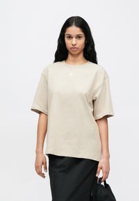 Bézs pamut póló, bővebb szabással, kerek nyakkivágással és rövid ujjakkal. A mellrészen kis fehér logó látható. Fekete szoknyával párosítva.