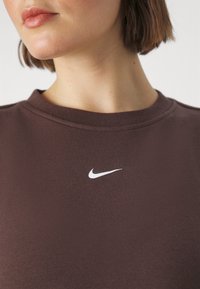 Donna che indossa una maglietta Nike marrone con un piccolo logo swoosh bianco centrato sotto il colletto.