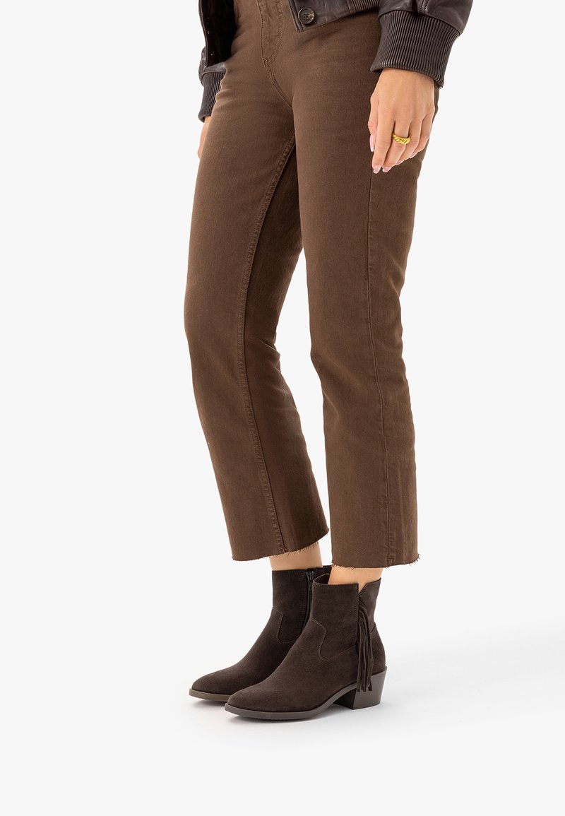 Femme portant un pantalon court marron et des bottines en daim marron foncé avec des pompons, debout sur un fond blanc.