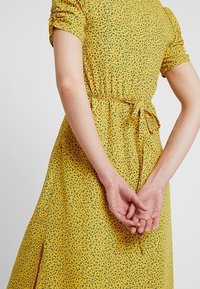 Persona che indossa un abito floreale giallo con maniche arricciate e una cintura annodata in vita, con le mani unite dietro la schiena.