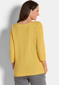 Gelber Strickpullover mit einem strukturierten Rautenmuster, Dreiviertelärmel und weitem Ausschnitt, kombiniert mit grauen Hosen.