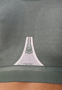 Sostén deportivo verde con canalé que presenta una etiqueta de logo blanca con un diseño texturizado. La tela parece elástica y suave, con costuras bien terminadas.