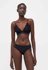 Bralette nera in pizzo con bordi ondulati e fascia con logo; slip neri senza cuciture abbinati con vita ondulata; texture liscia.