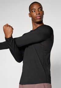 Langärmliges schwarzes Sportshirt mit rundem Ausschnitt, figurbetontem Design, glatter Textur und flachen Nähten, das von einer Person in einer Dehnpose getragen wird.