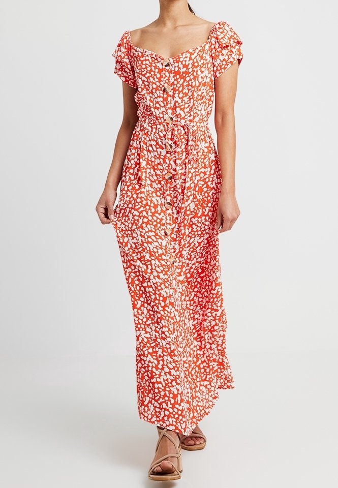 Femme portant une robe maxi orange à pois blancs avec manches courtes, boutons à l'avant et ceinture à la taille, associée à des sandales beiges.