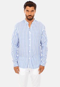 Camicia a righe blu e bianche a maniche lunghe con colletto alla coreana, chiusura con bottoni e polsini a contrasto. Indossata con pantaloni bianchi a vestibilità dritta.