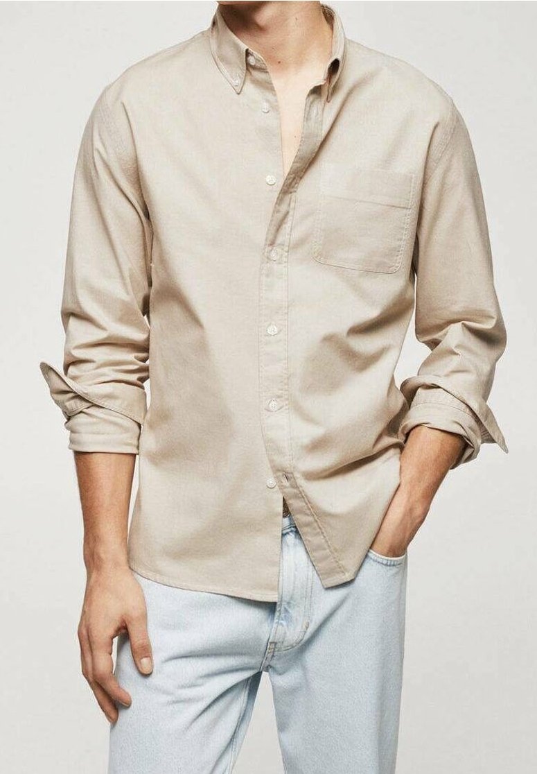Homme portant une chemise beige à boutons avec manches retroussées et un jean bleu clair, une main dans la poche, sur un fond uni.