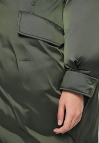 Doudoune verte avec une texture lisse et brillante. Dispose d'un poignet à boutons et d'une poche supplémentaire sur la manche pour plus d'utilité.