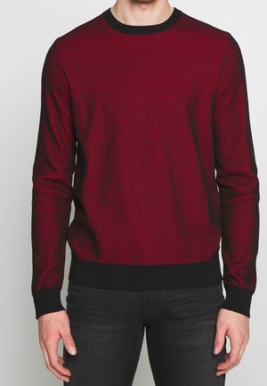 Man wearing a red and black textured sweater with black cuffs and hem paired with dark pants, standing against a plain background.