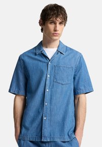 Camicia di denim azzurro chiaro a maniche corte con colletto a punta, bottoni frontali e una tasca applicata. Texture liscia e vestibilità rilassata.