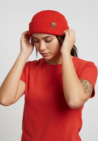 Red ribbed knit beanie with a folded cuff and a leather patch logo. Worn by a person in a matching red t-shirt.